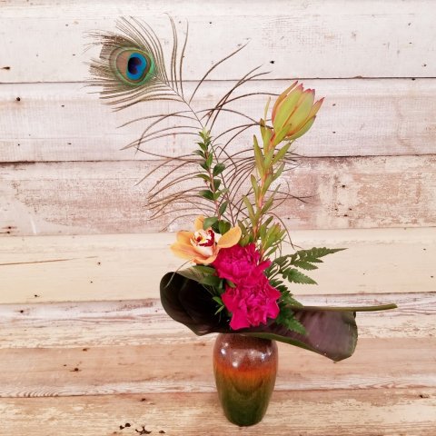 Modern/Contemporary Floral Design - Hot Pink Carnations, Orange Cymbidium Orchids with Green Luecadendron, assorted Greenery and a Peacock Feather. In a small Ceramic Container. Ceramic Colouring is a gradient from Green to Orange