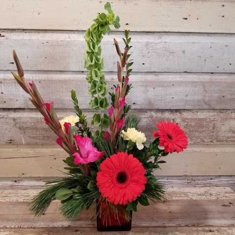 Flower arrangement - Red Gerber Daisies, Pink Gladiolas, Yellow Carnations, Bell of Ireland with aasorted greenery in a red Glass cube container